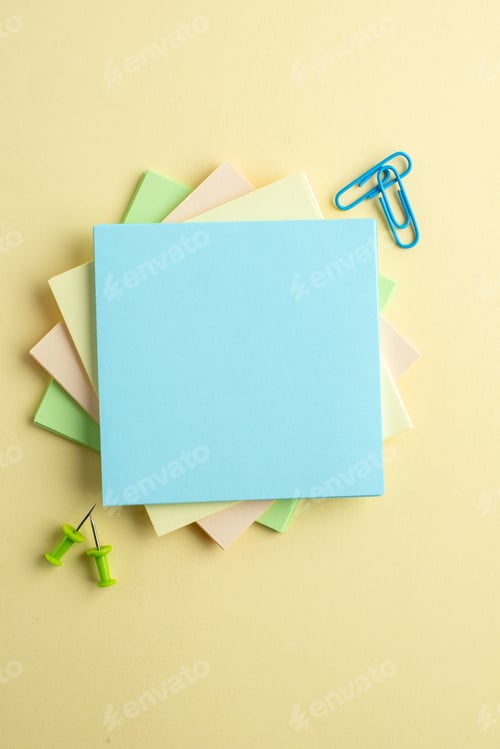 Preview: Vertical view of stacked coloured note cubes paper clips drawing pins on yellow background with