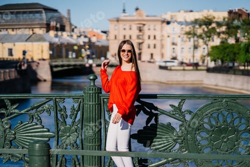 Preview: Attractive brunette young woman in red sweater, white pants walking on bridge at old town smiles