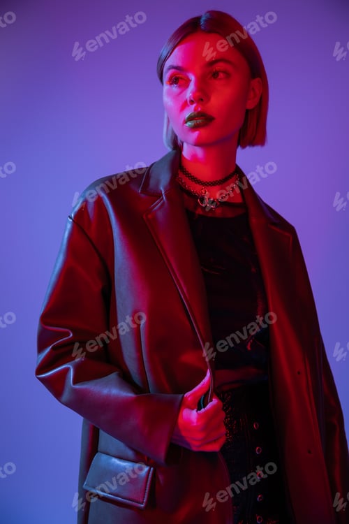 Preview: trendy woman in leather jacket and necklaces looking away in red light on blue background