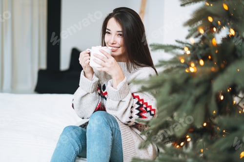 Preview: Young asian woman with cup of cocoa with marshmallow in room with Christmas tree at home
