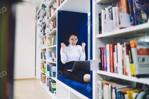 Preview: Happy ethnic woman using laptop while sitting on bench in library