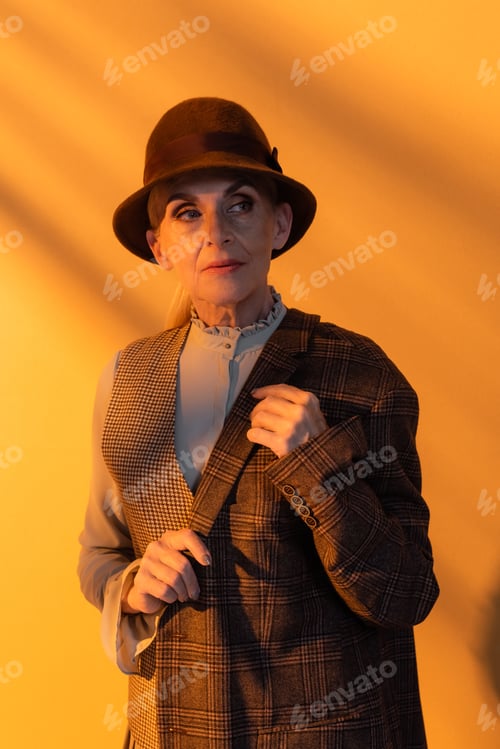 Preview: Senior woman in vintage clothes and hat holding jacket on orange background