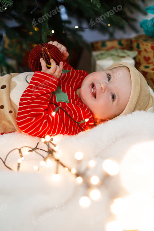 Preview: Cute happy baby in christmas costume playing with red bauble and lying under christmas tree