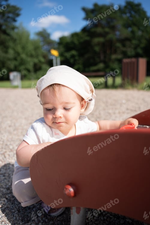 Preview: Baby in hat explores playground under blue sky, surrounded by greenery in joyful sunlight scene.