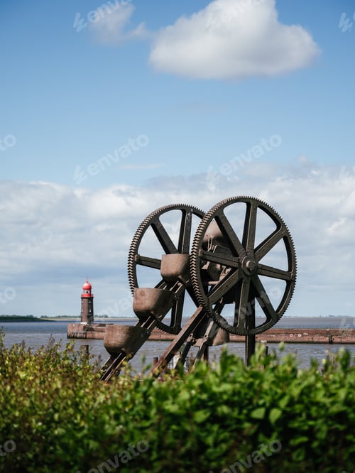 Preview: Majestic industrial wheel stands tall in the foreground in front of a picturesque lighthouse.
