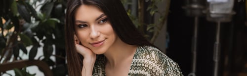 Preview: cheerful and dreamy young woman with long brunette hair and makeup holding hand near face