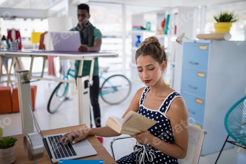 Preview: Female executive working on laptop while reading a book at desk