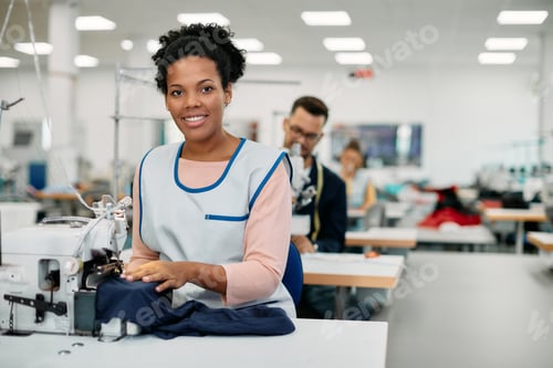 Preview: Young black seamstress working on sewing machine in textile factory and looking at camera.