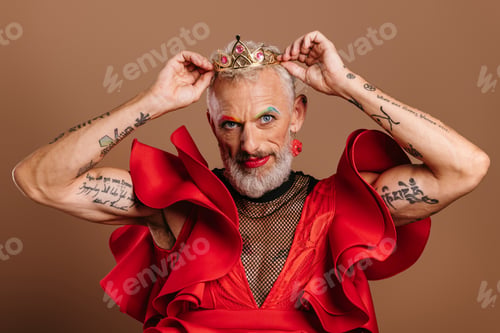 Preview: Portrait of confident mature gay man in beautiful red dress wearing crown against brown background