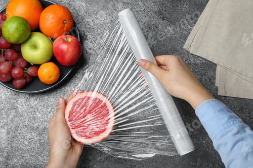 Preview: Woman putting plastic food wrap over half of grapefruit at grey table, top view