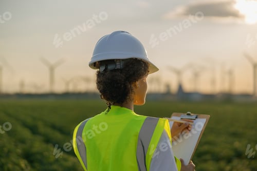 Preview: Back view of woman engineer in white helmet working with clipboard at renewable energy farm