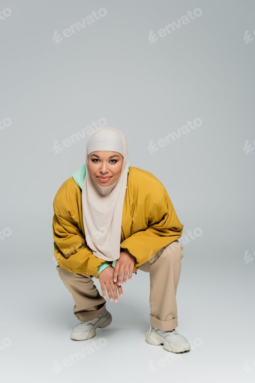 Preview: full length of fashionable multiracial woman in yellow bomber jacket and muslim hijab sitting on