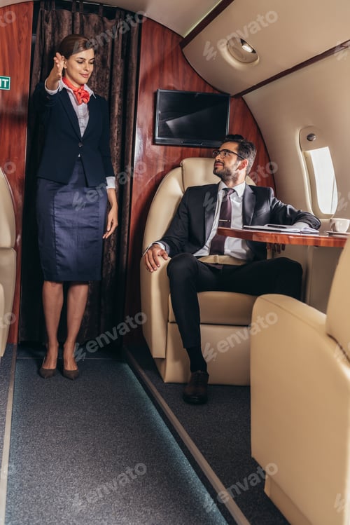Preview: flight attendant showing gesture to handsome businessman in suit in private plane