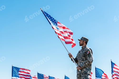 Preview: handsome military man in uniform holding american flag while standing against blue sky