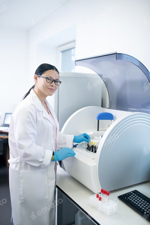 Preview: Dark-haired doctor in a white robe standing near hematological analyzer