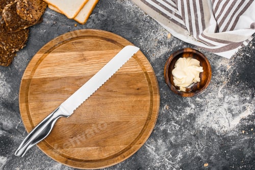 Preview: top view of wooden cutting board with knife and sliced homemade bread with butter in bowl on black