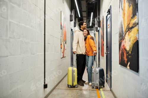 Preview: Relaxed multicultural couple with luggage embracing and standing together in a hostel hallway