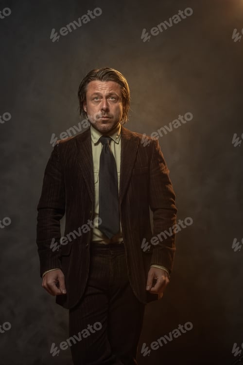 Preview: Cool Middle Aged Businessman In Dark Brown Suit And Black Tie In Front Of Grey Wall. Low Key Studio