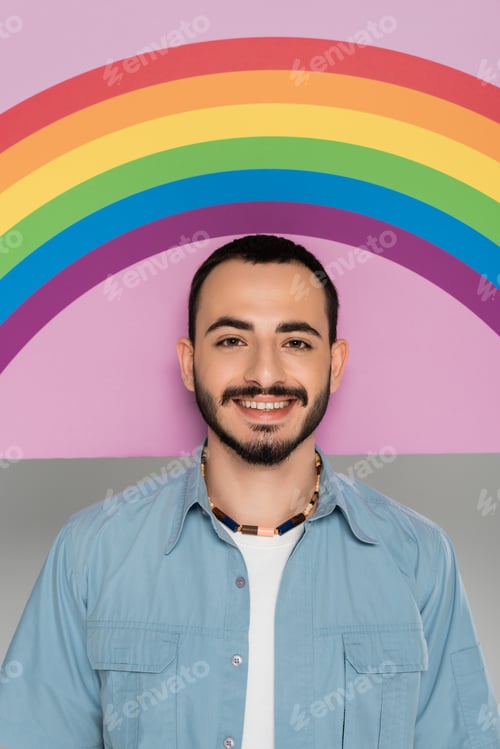 Preview: Portrait of smiling gay man looking at camera near placard with lgbt flag isolated on grey