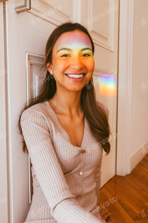 Preview: Young cheerful woman sitting on floor with rainbow light on her face