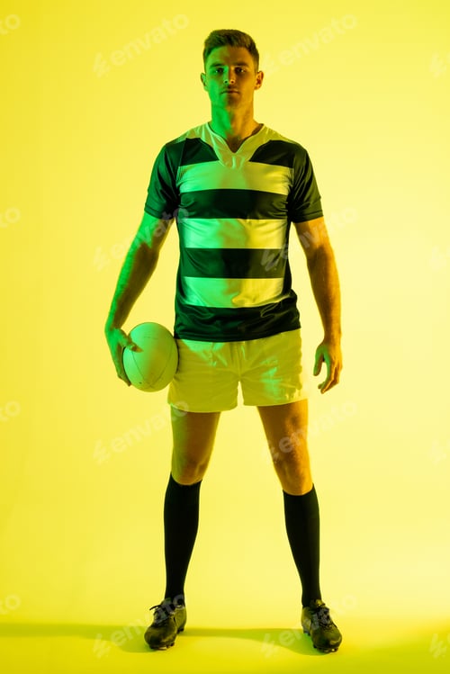 Preview: Portrait of caucasian male rugby player with rugby ball over yellow lighting