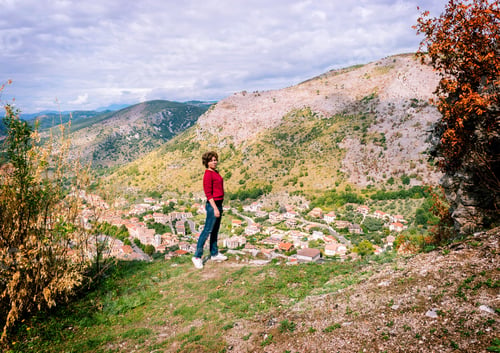 Preview: A traveler girl in a raspberry sweatshirt and jeans stands on a mountain above the Italian town