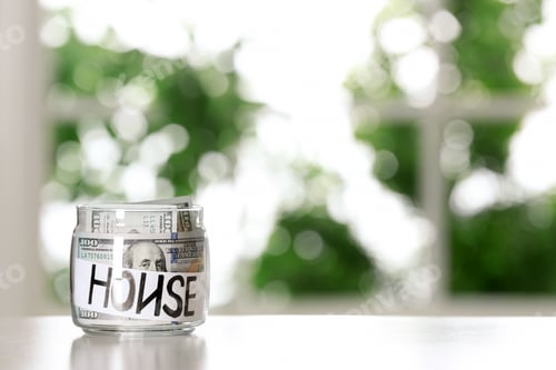 Preview: Glass jar with money and word HOUSE on table against blurred background, space for text