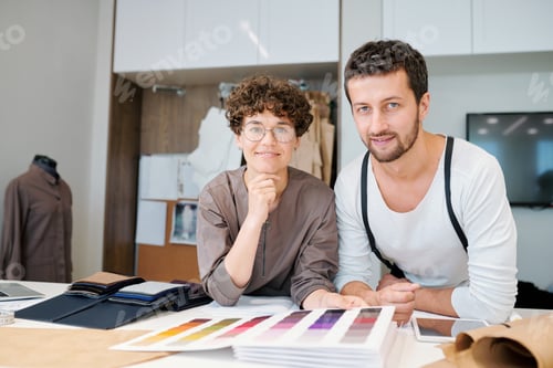 Preview: Young friendly fashion designers looking at you while bending over desk