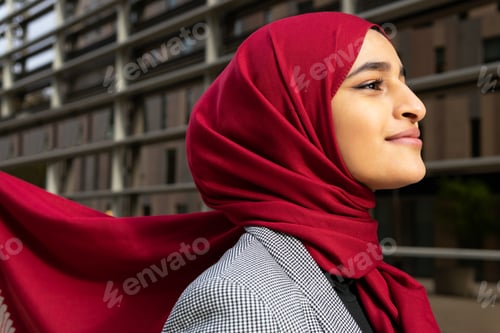 Preview: Side view of young content Arab female wearing red hijab standing on street and looking away