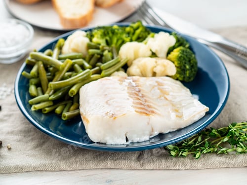 Preview: Boiled sea fish cod fillet with vegetables on blue plate, grey wooden table, side view. Healthy diet