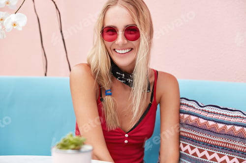 Preview: Cheerful female model has glad expression, wears red sunglasses, has blonde hair, sits on comfortabl