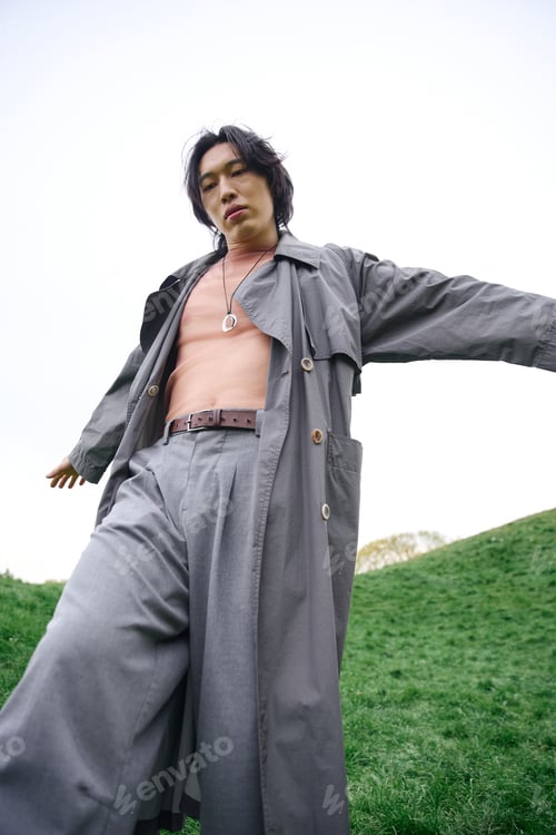 Preview: Young man showcases stylish trench coat in serene park setting with green backdrop
