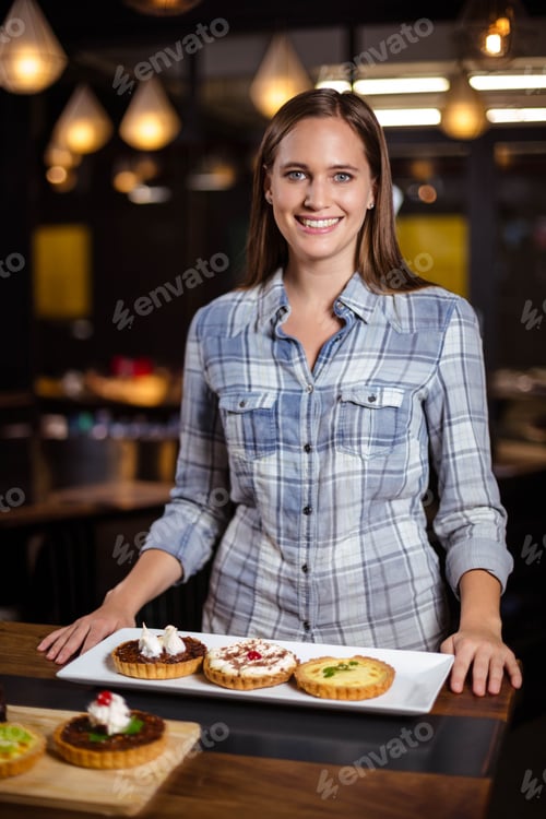 Preview: Adult woman in plaid shirt standing behind counter positioning platter with three tarts in bakery