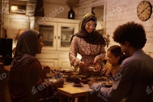 Preview: Young Muslim woman serving food to her family at dining table.