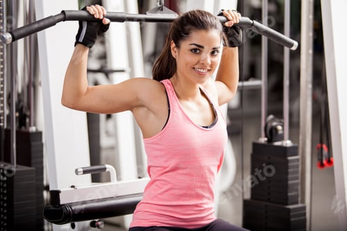 Preview: Young woman exercising back muscles using lat pulldown machine in gym