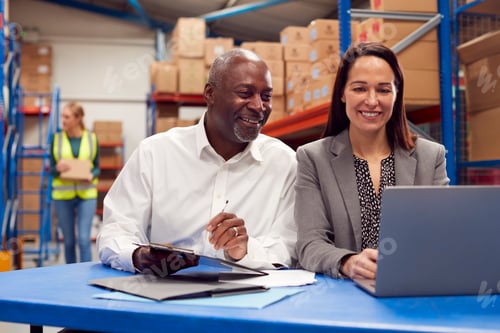 Preview: Male And Female Team Leaders Working On Laptop In Warehouse With Workers In Background