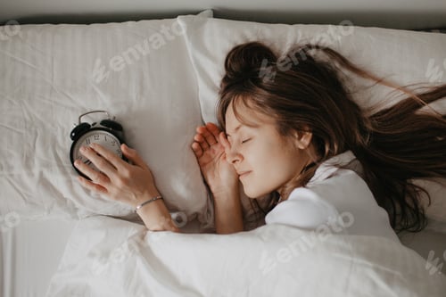 Preview: Pretty young woman with long hear sleeping in bed with alarm clock