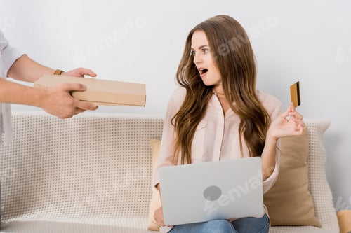 Preview: partial view of man with pizza in paper box and shocked woman with credit card and laptop at home,