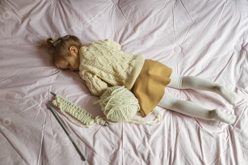 Preview: Happy little girl with blonde hair lying on the bed with wool skein for knitting hobby time bedroom