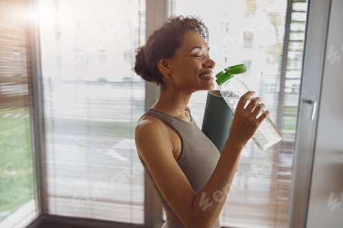 Preview: Smiling afro american woman drink water and holding rolled fitness mat after working out at