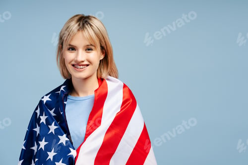 Preview: Smiling beautiful young woman with blonde hair wrapped in American flag looking at camera