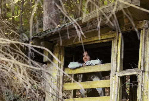 Preview: A young woman looks out the window of an abandoned house in the woods
