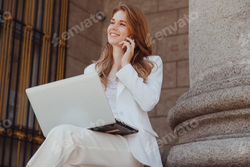 Preview: cheerful lady in white suit with laptop on knees speaking on mobile phone
