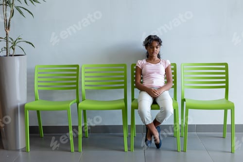 Preview: Bored mixed race girl sitting on green chair in hospital waiting room