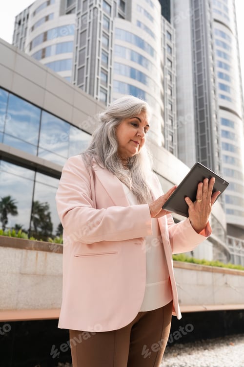 Preview: Senior businesswoman with long gray hair using a digital tablet in front of modern office buildings