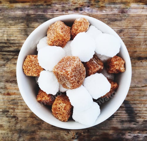 Preview: Looking down from above onto a bowlful of brown and white sugar cubes on a rustic wooden background