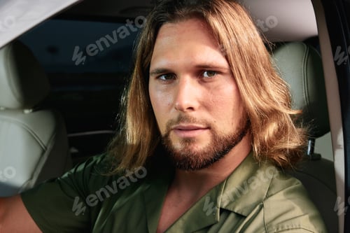 Preview: Man with long hair sitting in a car, looking confidently at the camera with a calm expression The