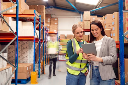 Preview: Manager Using Digital Tablet With Female Intern Inside Busy Warehouse Facility