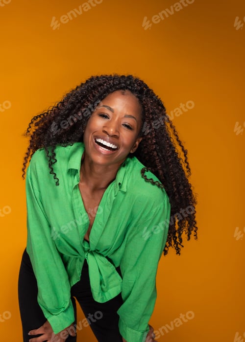 Preview: Cheerful African American woman in green shirt with curly hair laughing on yellow studio background