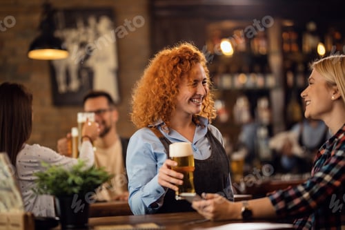 Preview: Happy waitress talking to a woman while giving her a glass of beer in a pub.